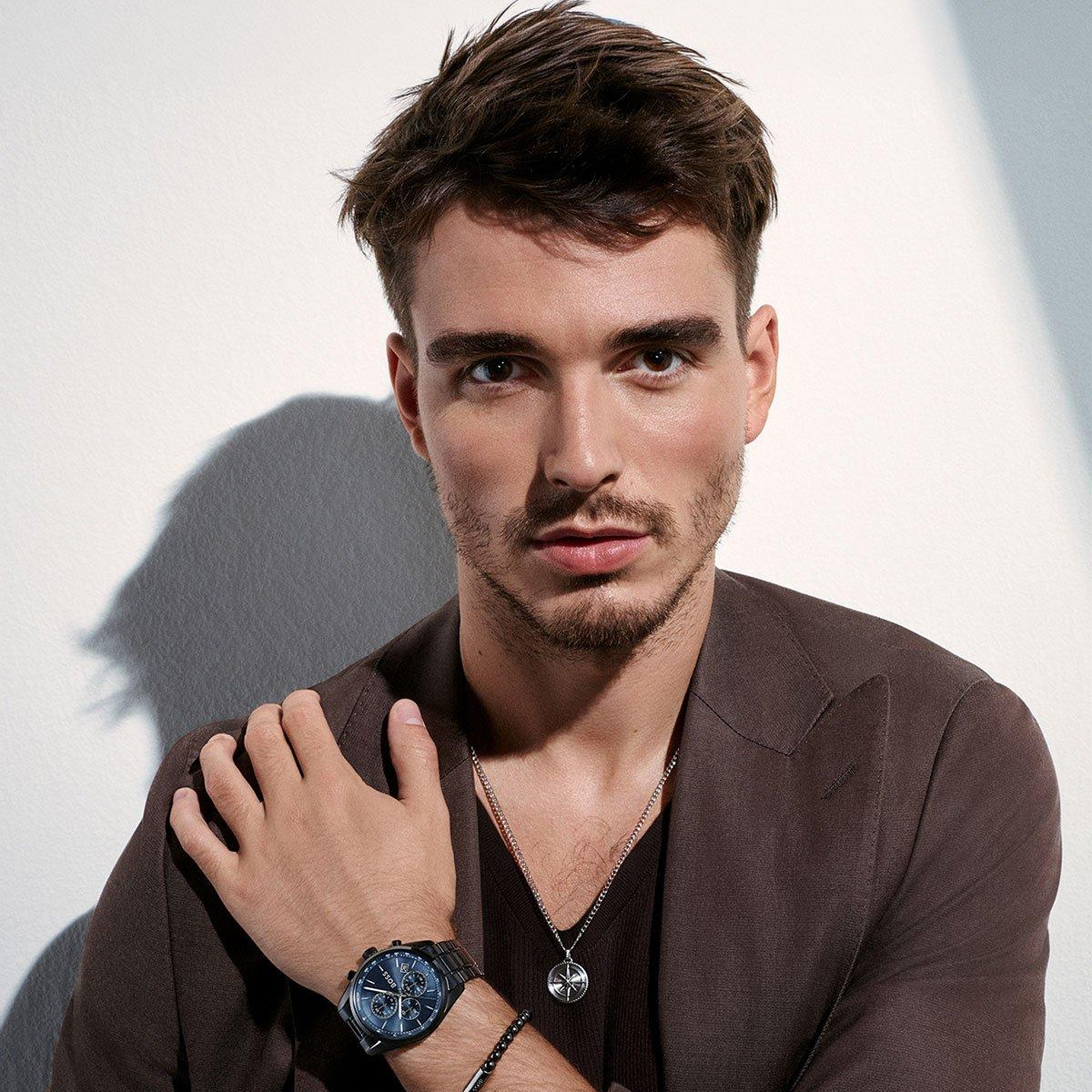 A man wearing a dark brown blazer, a silver pendant necklace, and a blue watch with a black strap, posing against a white wall with dramatic diagonal shadows.