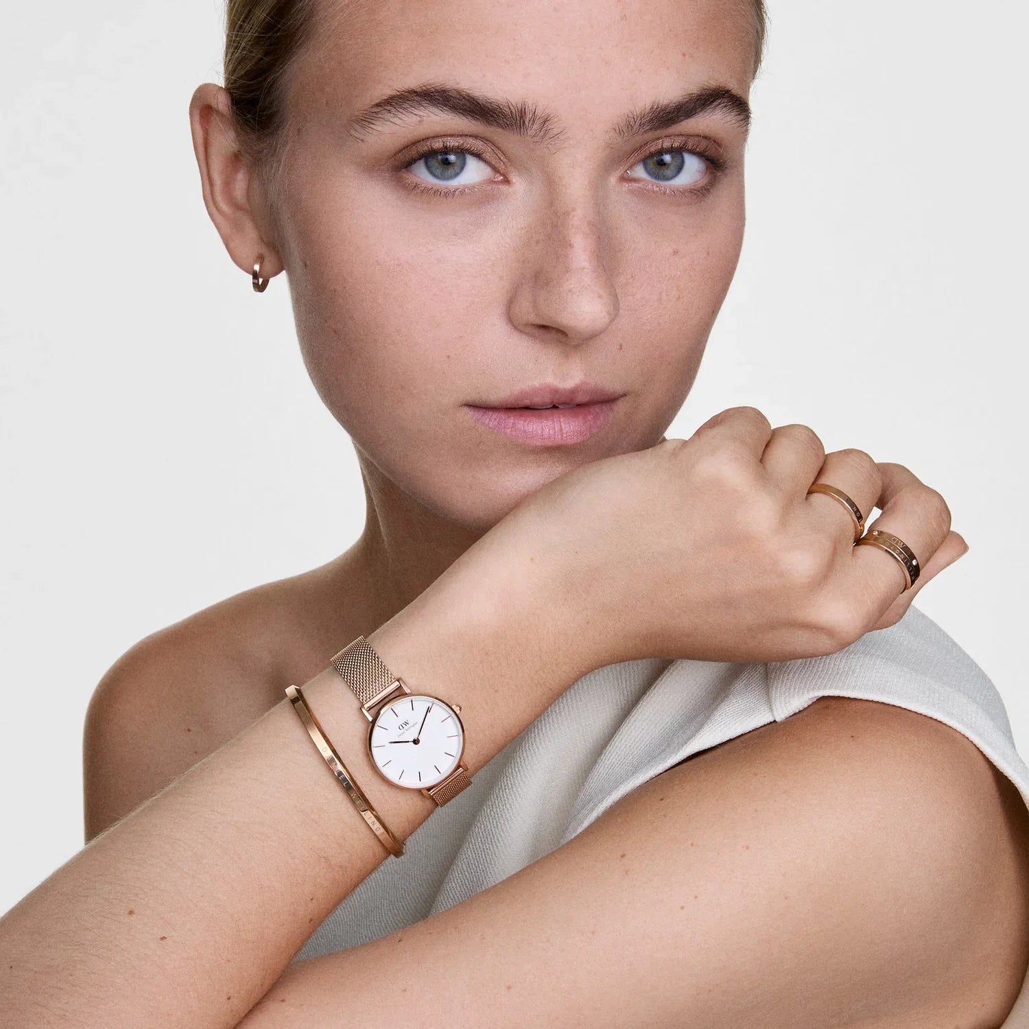 Woman wearing a rose gold watch and multiple rings on a plain background